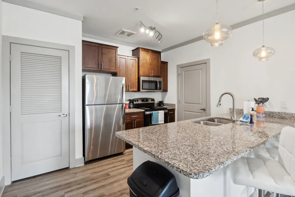A bright, open kitchen scene with granite countertops, stainless steel appliances, and modern cabinetry. Pendant lights illuminate the workspace, and a dining area with comfortable seating is visible in the background, suggesting seamless flow between cooking and dining.