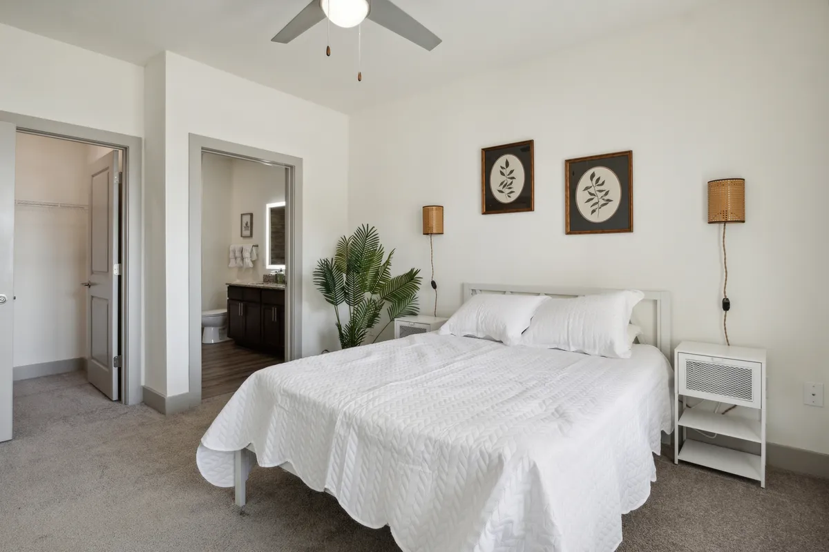 A tranquil bedroom featuring a bed with crisp white bedding, botanical wall art above the headboard, a lush green plant near the bed, and a nightstand with a lamp. Natural light streams in gently, creating an inviting and restful ambiance.