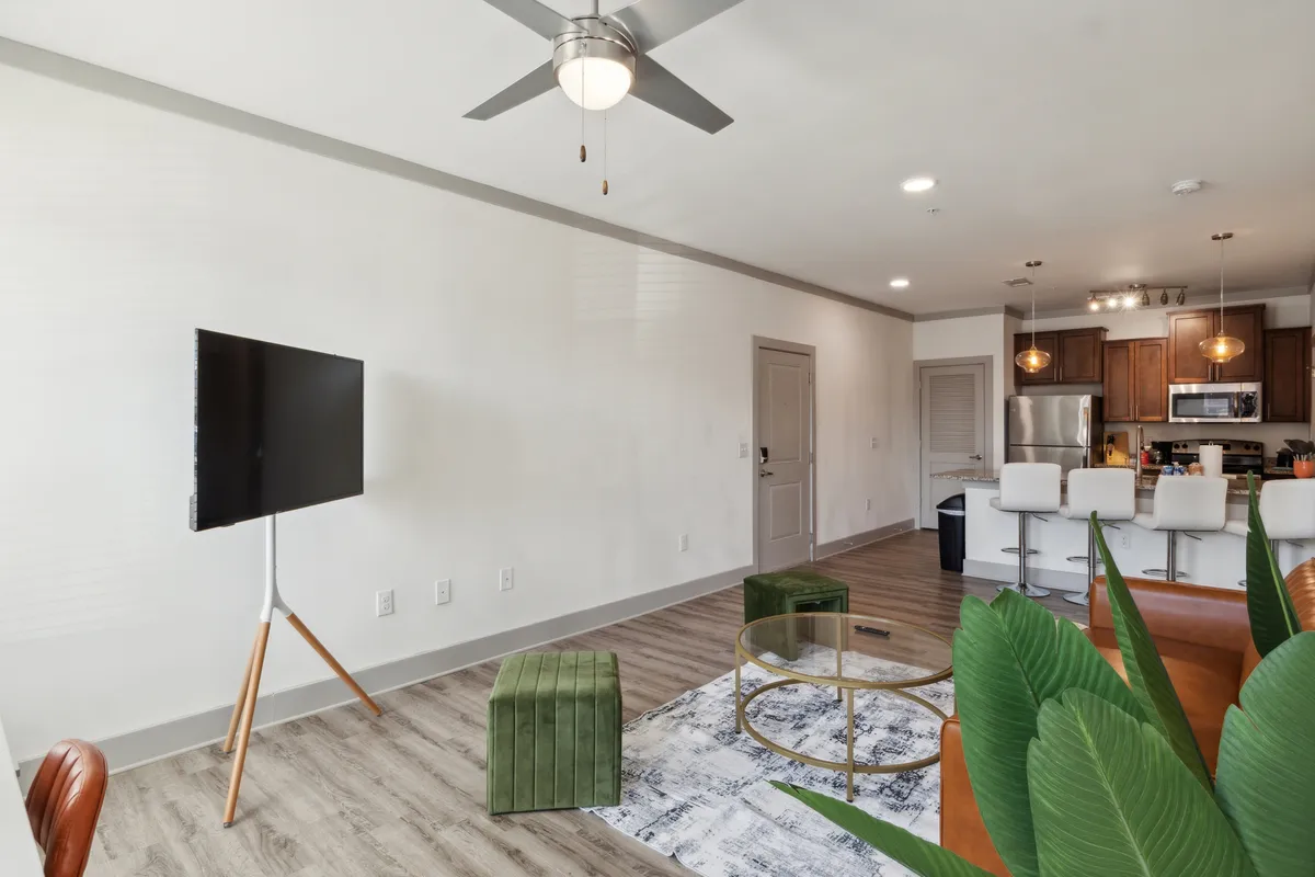 A bright, modern living area highlighting a TV on a stand, green ottomans, a sleek coffee table, and a partial view of the kitchen. The arrangement suggests a comfortable, tech-ready environment for streaming, socializing, or unwinding alone.