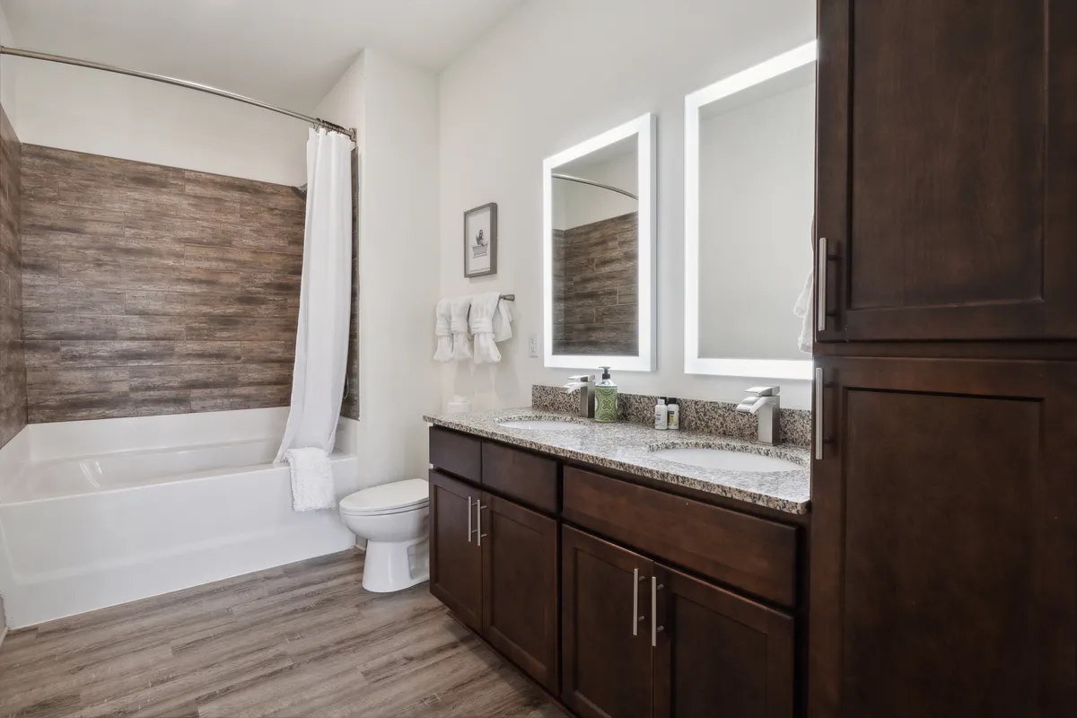 A modern bathroom featuring a spacious double vanity with a granite countertop, sleek mirrors, a bathtub with a shower curtain, and wood-like flooring. The space is bright, clean, and evokes a hotel-inspired atmosphere.