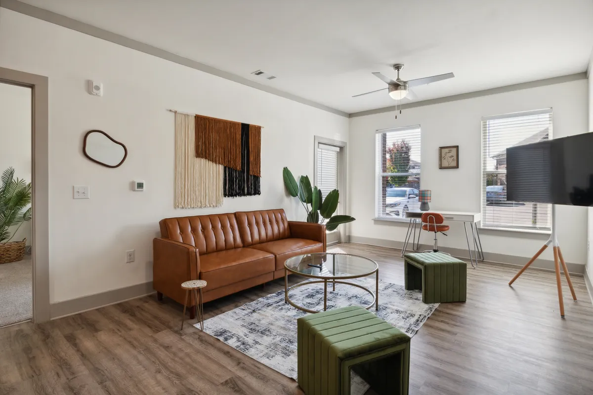 The image showcases a stylish living room featuring a plush brown leather sofa, a vibrant and colorful wall hanging, a sleek glass coffee table, and two green accent stools. The space is modern yet cozy, designed for relaxation or entertaining, with a TV stand completing the inviting setup.