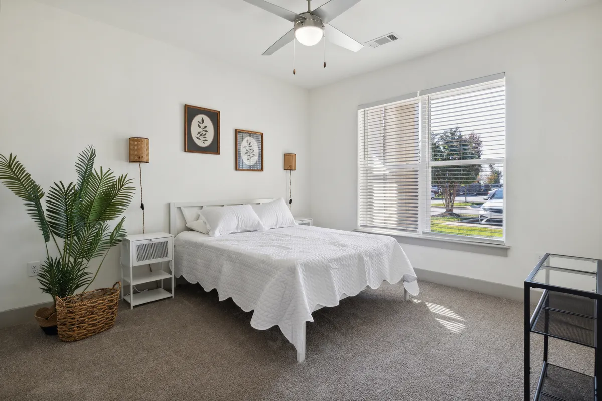 A cozy bedroom with a comfortable bed facing a window, flanked by houseplants and modern artwork. Sunlight brightens the room, highlighting its peaceful atmosphere and inviting space for relaxation or reading.