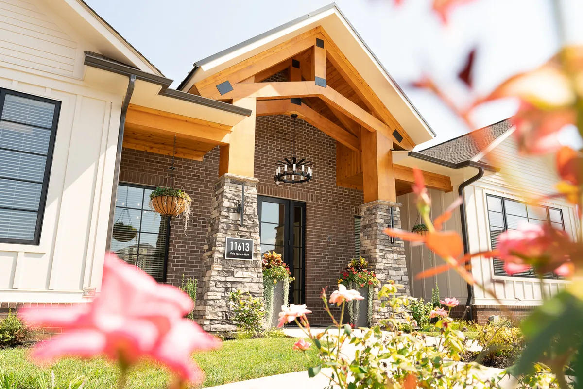A warm, welcoming entrance with wooden beams, a stone façade, decorative lighting, and vibrant flower beds. The scene suggests hospitality and curb appeal, inviting residents and guests to step into a stylish living space after a day in the city.