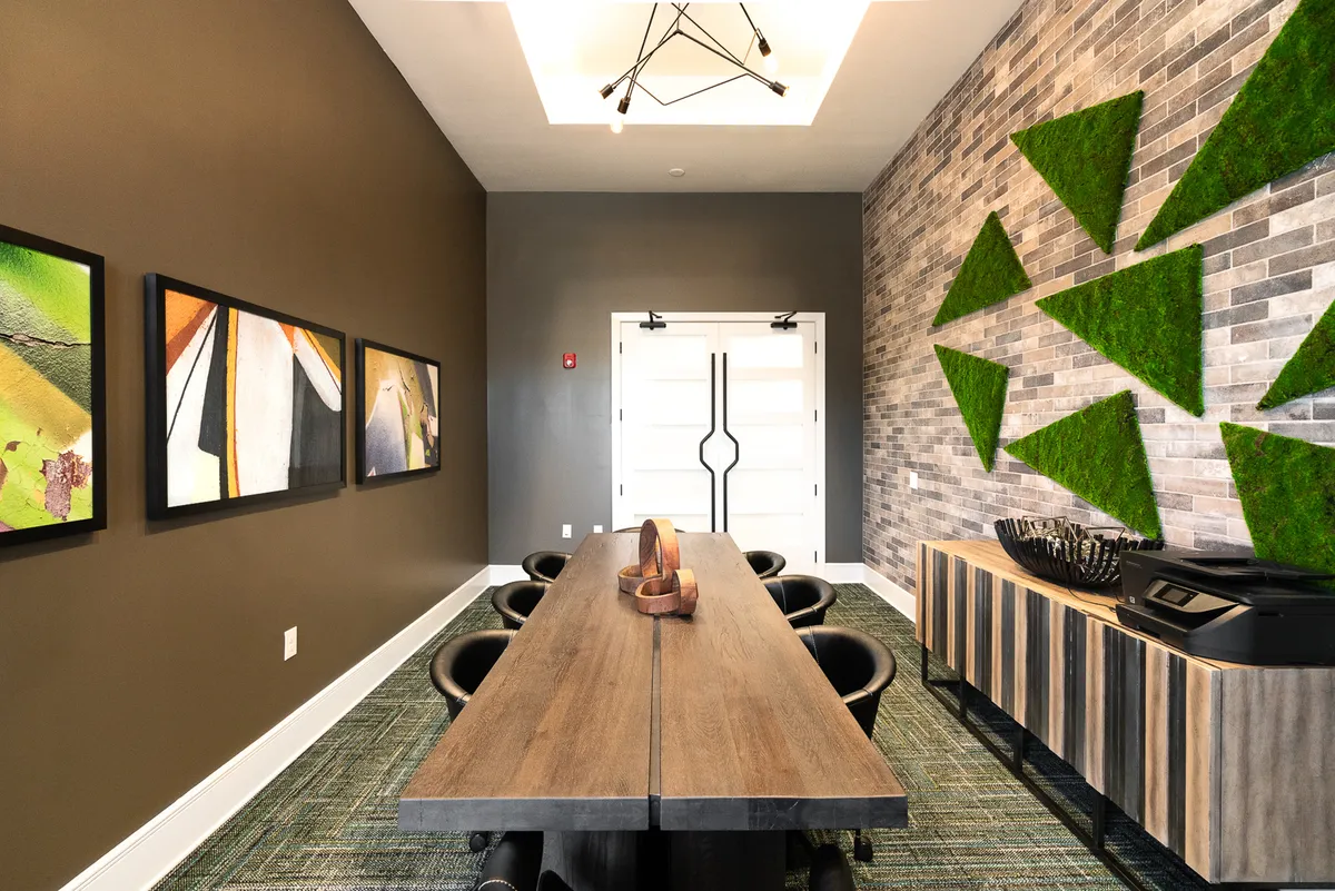 This image features a long wooden table surrounded by modern seating, highlighted by geometric wall art and a decorative moss wall. The space is illuminated by a stylish light fixture, blending professionalism with comfort in an open, communal area.