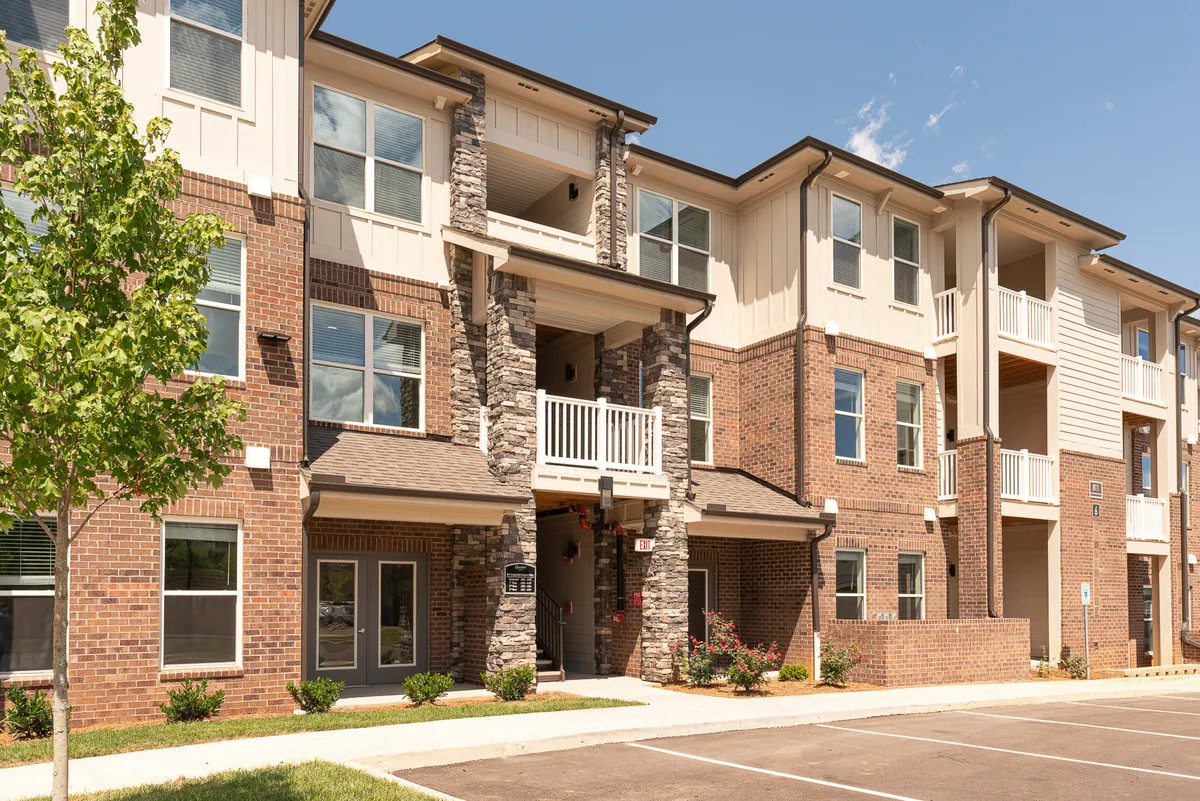 A broad view of the building's stone facade, featuring a sunny balcony, manicured landscaping, and a welcoming entrance. The courtyard area hints at communal gatherings and provides a glimpse into the property's connection to the outdoors and city life.