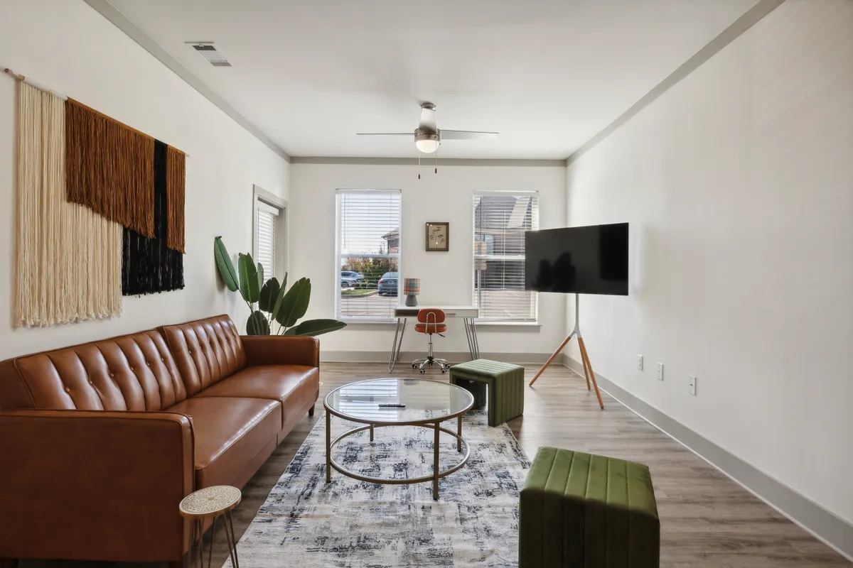 This image showcases a chic living area featuring a brown leather sofa, a round glass coffee table, a textured wall hanging, and green accent stools. A TV sits on a modern stand, and the room is styled with inviting decor and soft lighting that exudes comfort and sophistication.