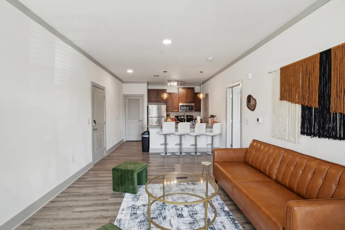 A modern living space with a brown leather sofa, glass coffee table, green ottoman, and a view toward the open kitchen area. The spacious layout encourages socializing and entertaining, while maintaining a warm and inviting atmosphere.